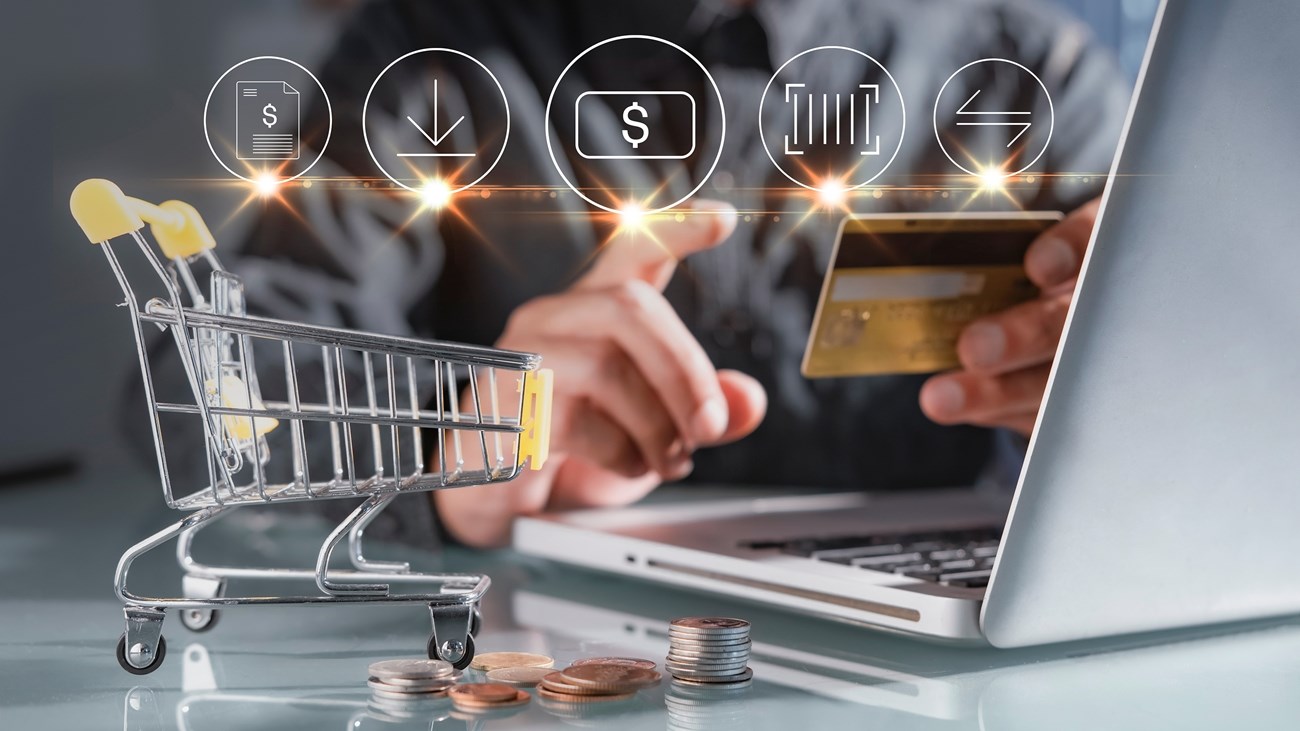 Person making secure online payment with credit card on laptop, miniature shopping cart and coins on desk with e-commerce icons