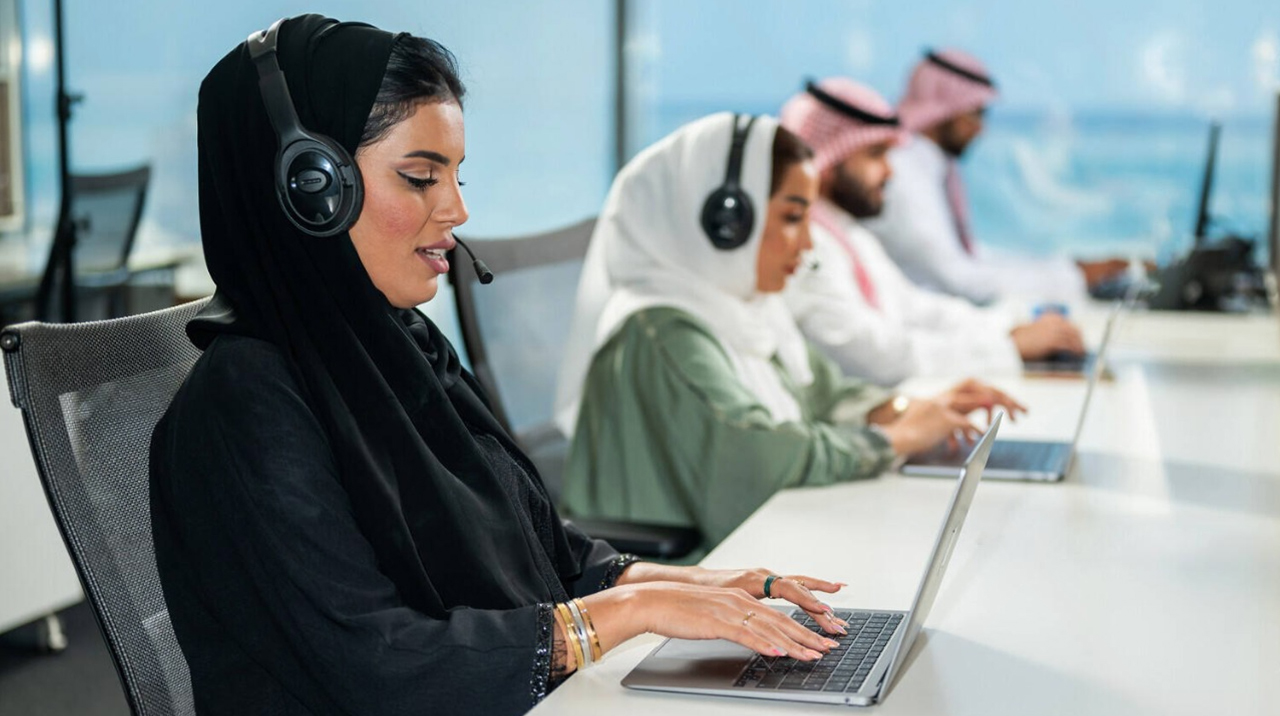 Saudi customer support team in traditional attire wearing headsets, working on laptops at modern managed services help desk
